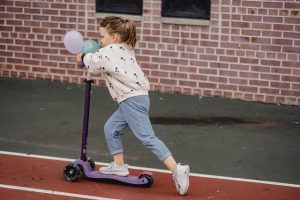 A kid is using a scooter, kicking the ground while holding balloons in her hand.