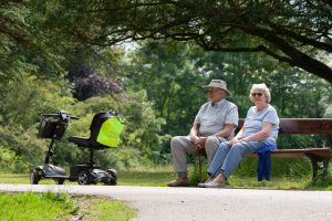 Two elderly people sit on a bench with a scooter. 