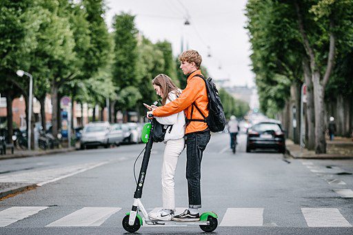 A person in tandem observing scooter safety while they’re riding across the street, going to school. 