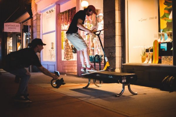 A teen boy doing scooter tricks on a bench at night while his friend films and shines a light on him