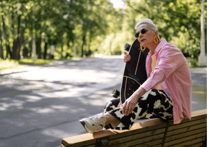 An elderly woman exudes coolness as she rests on a bench with her skateboard, proving that skateboarding knows no age. Her stylish attire and sunglasses reflect a vibrant spirit, embracing the culture of skateboards with ease and flair.