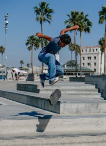 A skateboarder is caught in the midst of a challenging jump over a set of concrete steps in a sun-drenched skatepark. Dressed in casual blue attire that matches the sky above, the individual's body language exudes concentration and athleticism. The scene is set against a backdrop of palm trees and a historic building, reflecting the urban environment often associated with the sport of skateboarding.