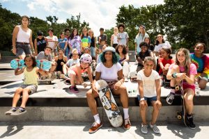 A vibrant group of young individuals stands together at a skatepark, representing a rich diversity of ethnicities and styles. They are positioned on various levels of skatepark terrain, with many holding skateboards and wearing helmets and protective gear. Their faces reflect the community and joy of skateboarding, with a clear, bright sky overhead and lush green trees surrounding the area, providing a perfect setting for their shared passion.