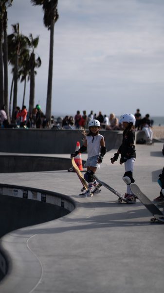 Two kids wearing safety gear while skateboard riding in a skatepark with other children. 