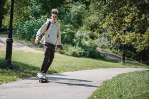 A teenager cruising down a park pathway on their skateboard, gripping another skateboard in their hand, with various skateboard likely being their passion as the teenager enjoy a sunny day skateboarding in nature. This man is having a great time riding in a cemented pathway with a good board.