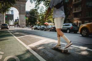 A young longboarder crossing the street on their skateboard, showcasing the urban commute on skateboard, with the backdrop of city traffic and a historic arch, highlighting the integration of skateboarding into city life. Take not though that safety is always a policy when riding.
