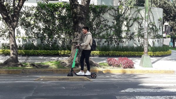 A man with a black pants and brown jacket rides his best scooter while commuting during a bright day. 