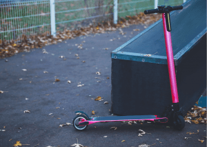 There is a black-pink scooter touching the black bench. There are also dry leaves that fell on the ground.