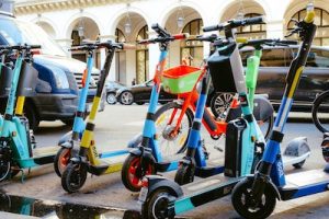 Scooters and Deck: A group of colorful electric scooters with their scooter decks visible, lined up on a city sidewalk. Some scooters have helmets hanging from the handlebars. The scooters and each deck is colroful. The scooters and each deck is flat. 