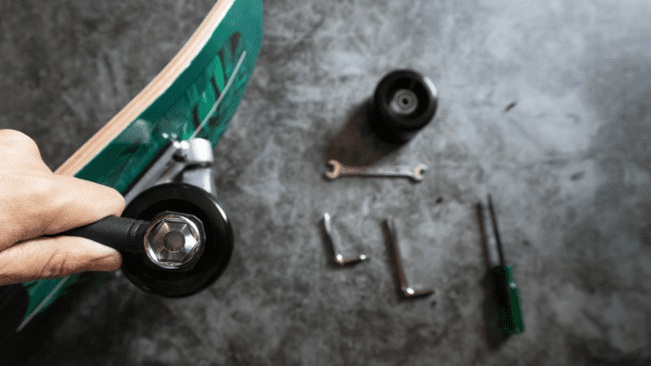 A hand holding a small wheel. On the floor are skate tools and a skateboard.