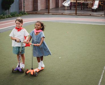 Two kids wearing white shoes ride their three-wheel scooters while playing outdoors.
