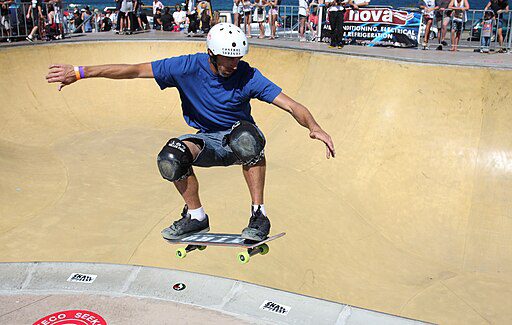 A skilled man in a vibrant blue shirt effortlessly executes a stylish skateboard trick in the skating area, showcasing impressive balance and control, surrounded by the energetic atmosphere.