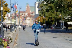 A bald man wearing shades is scooting on the road. 