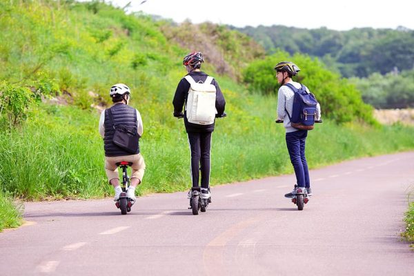 A boy with a sling bag rides his electric scooter with seat while other two boys ride their electric scooters without seats.