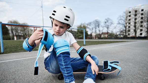 A kid looking at his skateboard safety equipment while sitting on his skateboard.