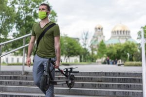 A man wearing a green mask and green shirt carries his scooter conveniently while walking. 