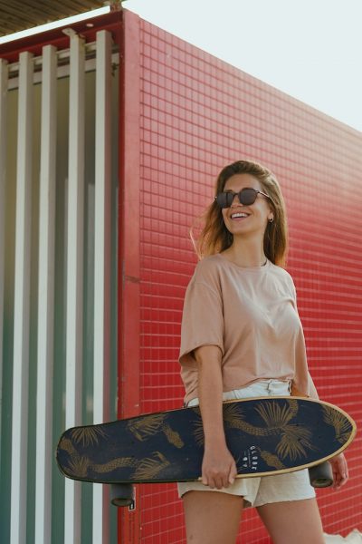 A woman wearing brown shirt carries her cruiser board while smiling. 