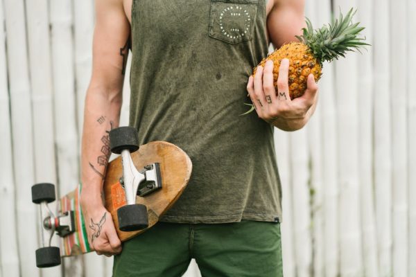  A man in a green shirt stands and holds a skateboard in one hand and a pineapple in the other.