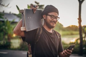 A skateboarder is lifting his electric skateboard while texting.