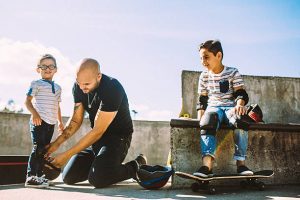 Dad helping his skater son wear skate gears before riding the skateboard at a public skate park.