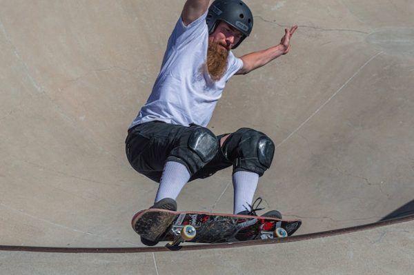 A skateboarder with a beard, wearing a helmet and knee pads, performing a trick in a skatepark.