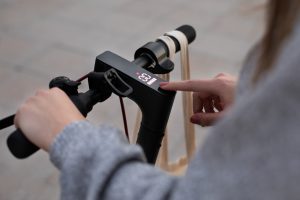 A woman wearing a gray sweater holds the handlebar of the scooter while pressing the app feature. 