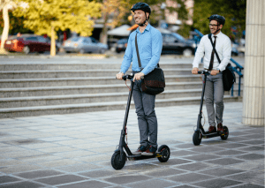 Two adult male are using scooters to commute to work. They are both wearing helmets as protective gears.