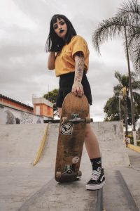 girl holding the board while posing for the camera wearing orange shirt and vans for her shoes, as well as riding in a short