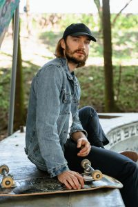 A man with beard wearing a black hat and blue jacket is holding his skateboard