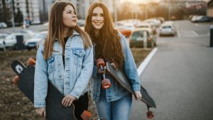 Two Smiling Women Heading to the Skateboarding Park while talking about the current popular culture around them, both of them seem to be having fun in their skate attire.