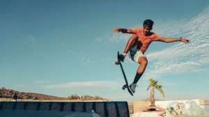 A young man wearing an orange shirt is performing some advanced skateboard tricks in broad daylight.