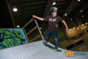 A boy wearing skateboard protective gear practices skateboard trick on skate ramp indoors