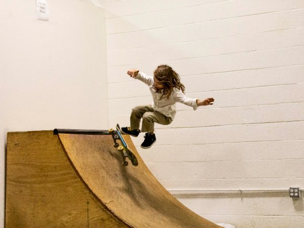 A woman with black shoes does skateboard tricks confidently in well-lit skate ramp indoors. 