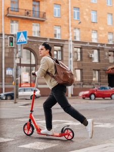 A man with a brown bag and black pants rides his kick scooter, one of the excellent kinds of scooters, while cautiously traveling outdoors.