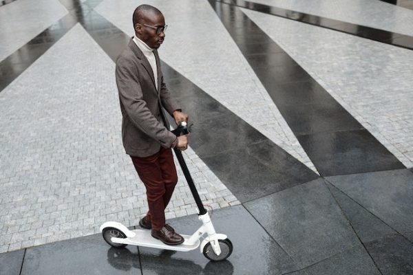 A brown man is riding his new replaced grip tape scooter in a hallway with a brown and white color combination of tiles.