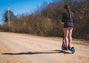 A woman with braided hair and a black shirt rides her scooter on a bright day.