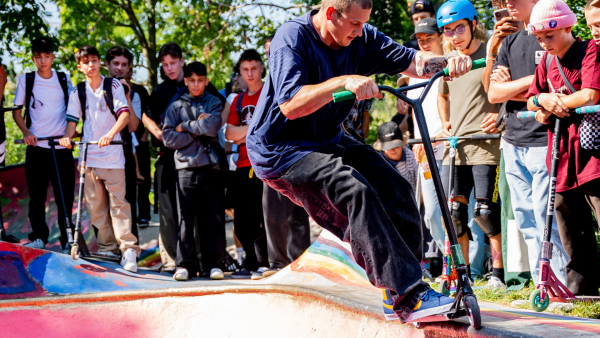 Scooter exhibitionist is performing the tricks with his best stunt scooter in front of a crowd of people and other scooter enthusiasts.