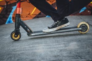 A man wearing black shoes and pants rides his stunt scooter with one foot up and prepares to show his best scooter tricks and maneuvers. 
