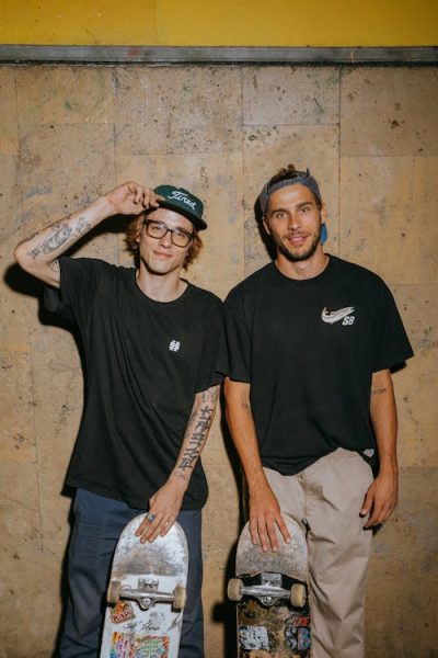 2 men wearing oversized t-shirts featuring slogans and logos. Skateboard apparel trend.
