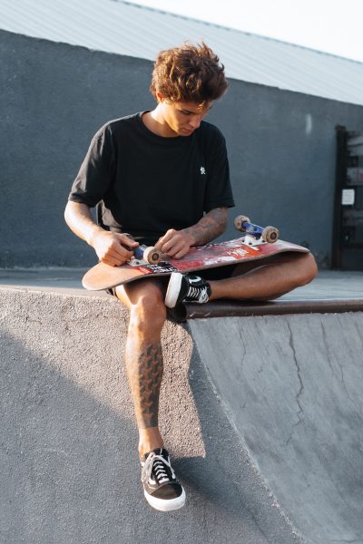 A skateboarder in black shirt doing some bearing maintenance on his skateboard. 