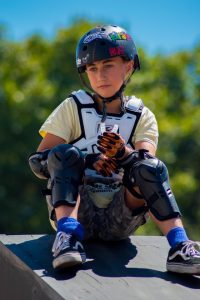 Young scooter enthusiast wearing protective gear and a helmet with Spiderman stickers, sitting on a ramp.