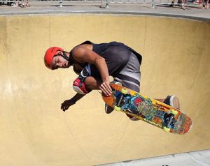 An intense moment of action is captured as a skateboarder, clad in dark attire and a vivid red helmet, carves deeply into the curve of a sandy-colored skatepark bowl. The skater's worn knee pads tell a story of dedication and frequent practice. This image evokes the adrenaline and technical skill inherent in the sport of skateboarding, with the smooth concrete surface of the bowl shining under the bright sunlight.