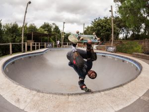 A skateboarder donned in protective gear, including knee pads and a dark helmet, is captured in a dramatic pose while skateboarding inside a deep concrete bowl. Their silhouette is sharply defined against the overcast sky, with the surrounding greenery of the park faintly visible. The intensity of the skater's focus and the action-packed atmosphere of the skatepark are palpable, reflecting the serious athleticism that skateboarding demands.