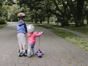 Two children stand with scooters on a park path. Each scooter's deck is distinct; one scooter's deck is adorned with purple accents. The scooters' deck gleams against the backdrop.
