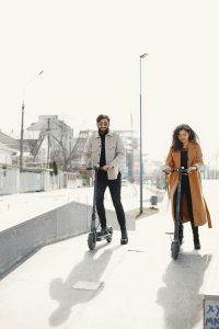 Scooter Decks: A man and a woman outdoors in a bright, urban environment. The man is riding an electric scooter, while the woman is riding a scooter beside him. The scooter's deck and the couple's relaxed demeanor suggest a leisurely activity. The scooter deck is black. Each scooter deck is flat.