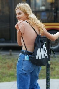 A blonde girl with a pink blouse and denim pants is smiling.