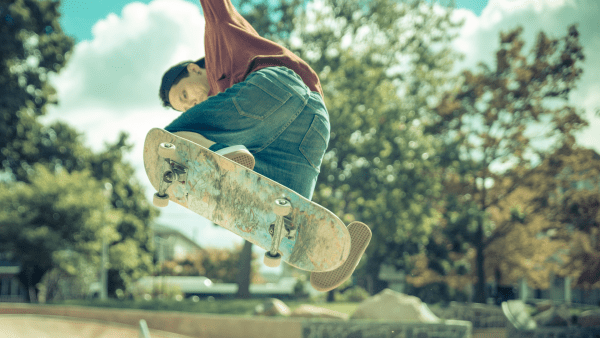 A dynamic moment is frozen in time as a skateboarder is captured mid-air against a clear blue sky, executing a skillful trick. The individual's intense focus and physical prowess are evident as they maneuver their well-worn skateboard with precision. The backdrop of the park is a lush landscape of trees and a hint of urban structures, highlighting the juxtaposition of nature and the urban skateboarding scene.