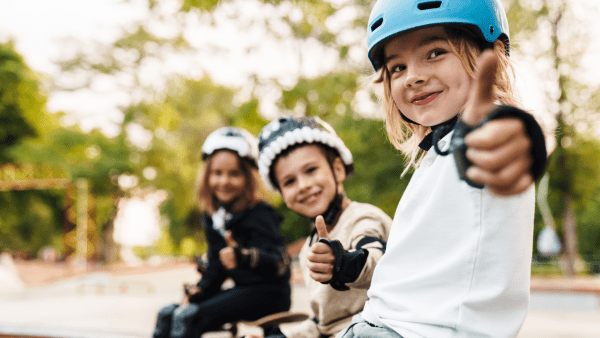 Three exuberant children are seated on the edge of a skate park, each giving a thumbs-up to the camera with beaming smiles. The child in the foreground wears a light blue skateboarding helmet and a white shirt, while the other two, slightly out of focus in the background, also sport protective skateboarding gear.