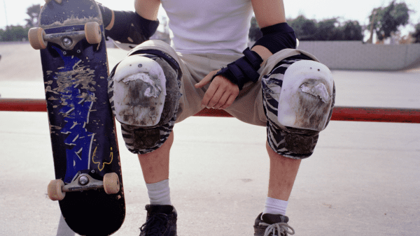 The image provides a close-up view of a skateboarder's lower half, with a focus on the heavily used board clutched in one hand and the battered knee pads that speak to many hours spent practicing. The textured concrete of the skatepark ground and the bright red rail in the background suggest a space where skills are honed and the skater's dedication to the craft of skateboarding is as evident as the scuffs on their protective gear. The stance is one of readiness and resilience, emblematic of a skateboarder's perseverance and the continuous journey of improvement and mastery in the sport.