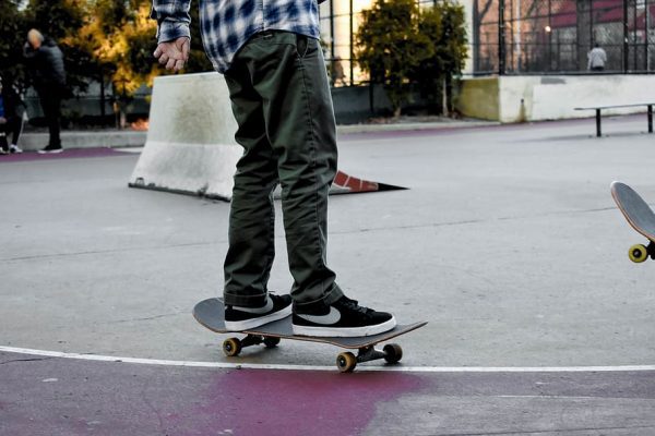 Person in custom green pants and a checkered shirt standing on a custom skateboard at a skatepark.